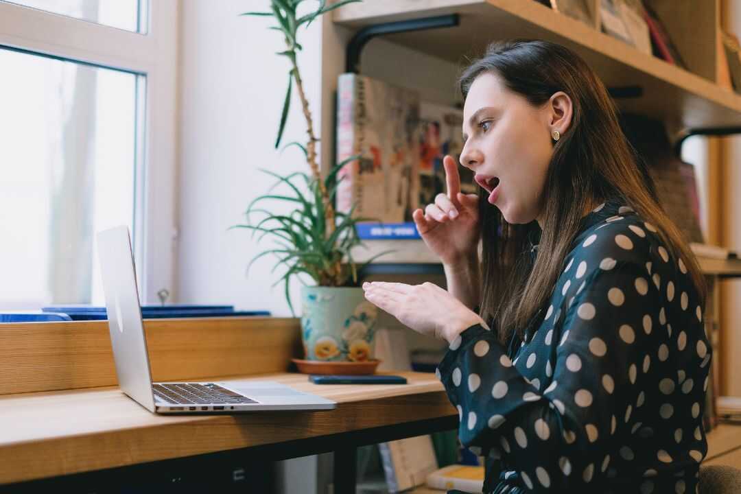 A woman in a polka dot blouse engages in a virtual meeting from a cozy home office, gesturing enthusiastically towards her laptop screen. This image represents team-building activities for HR professionals seeking to enhance collaboration and communication within their teams.