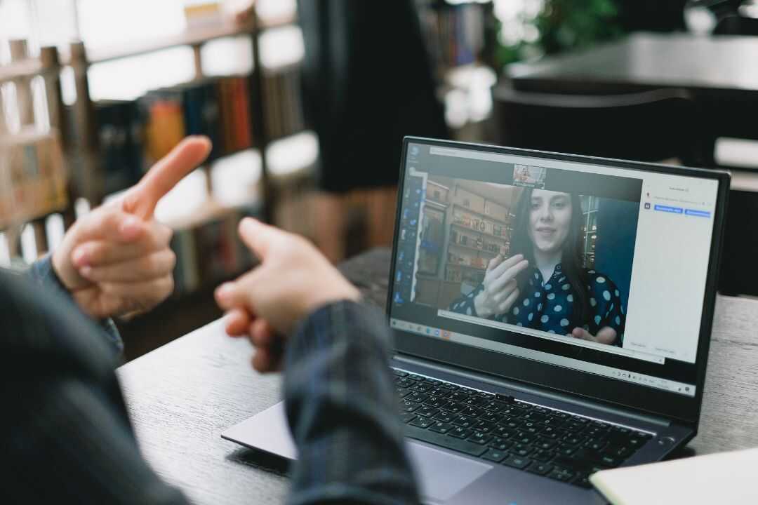 A person using a laptop to participate in a virtual team-building activity, with a colleague visible on the screen in a video call setting. The hands of another participant are seen gesturing, highlighting the interactive nature of online events designed for enhancing collaboration and communication in remote teams. Suitable for HR professionals seeking engaging team-building solutions.