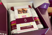 Box containing wine bottles wrapped in branded paper and an Urban Grape event flyer, highlighting a team-building wine-tasting experience for HR professionals.