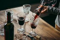 A person pours red wine into a crystal glass on a wooden table. The setting includes several filled glasses, an unopened wine bottle, and a corkscrew, suggesting a relaxed and elegant atmosphere for a team-building wine tasting event.