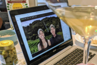 A laptop displaying a virtual meeting with two people against a vineyard backdrop, surrounded by snacks and a cocktail glass, suggesting a virtual team-building event.