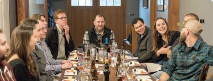 A group of people seated around a long table enjoying a team-building meal. They appear engaged and smiling, surrounded by plates of food and glasses, in a cozy dining setting.