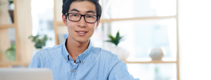 A person sitting at a desk in a well-lit office, holding a smartphone and smiling at the camera, with a laptop in front. The scene conveys a professional setting, suitable for HR professionals looking to book team-building events.