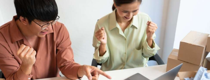 Two colleagues excitedly celebrate a successful team-building event booking on a laptop, surrounded by boxes and digital devices, suggesting an engaging and effective activity for HR professionals.